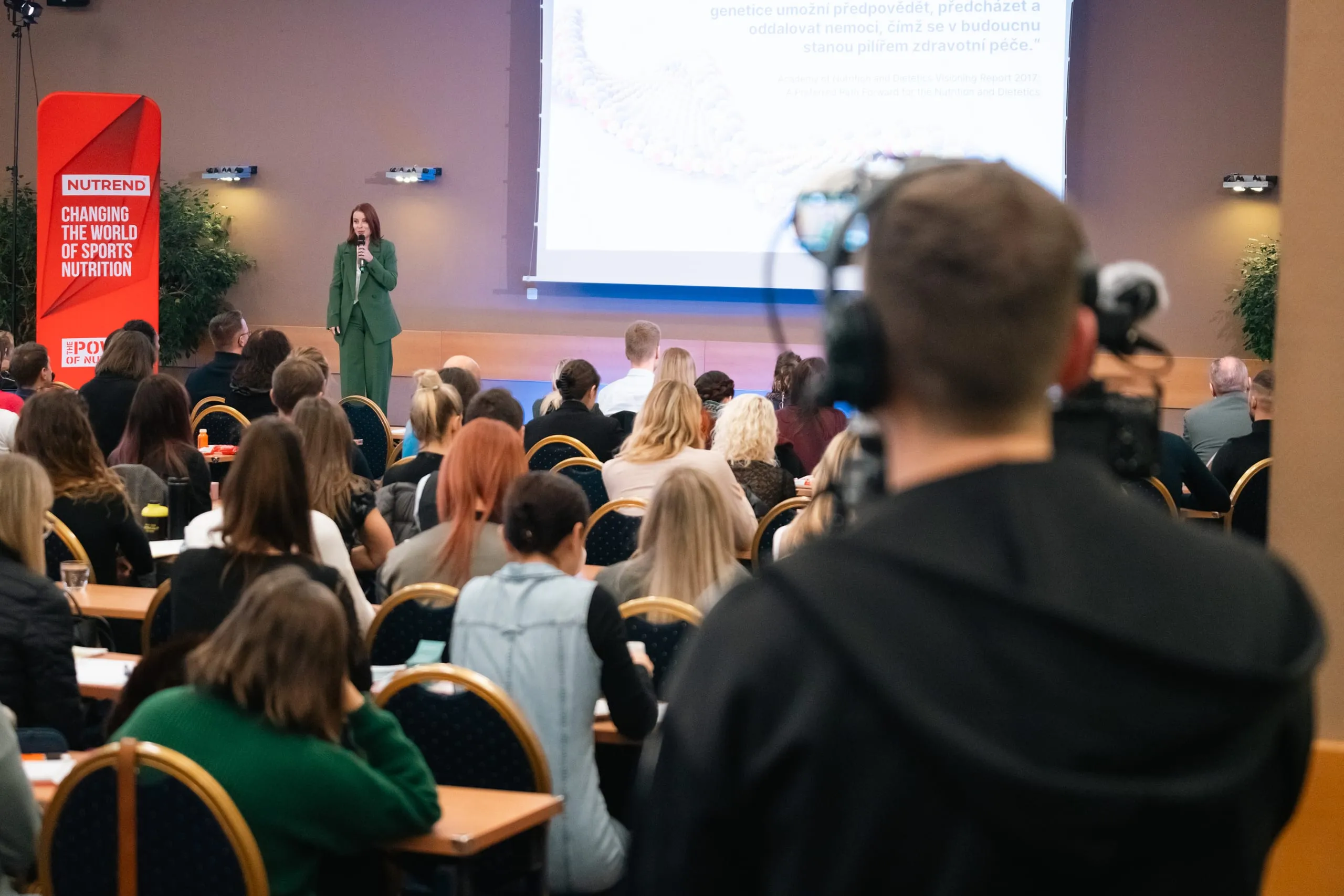 Řečnice u mikrofonu před zaplněným konferenčním sálem, vpravo kameraman Forpix Live